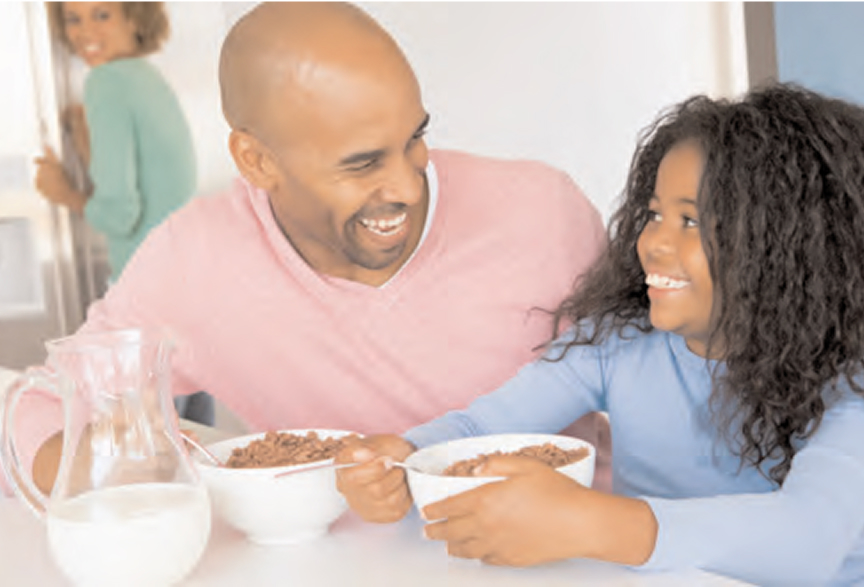 A man and a young girl smiling at each other while eating cereal at a table. A woman in the background is entering the room, smiling at them.