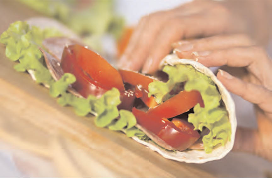 Hands preparing a wrap sandwich with lettuce and sliced tomatoes inside a soft tortilla