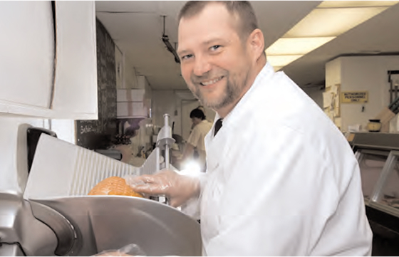 A smiling man in a white coat and gloves is slicing deli meat in a grocery store deli department.