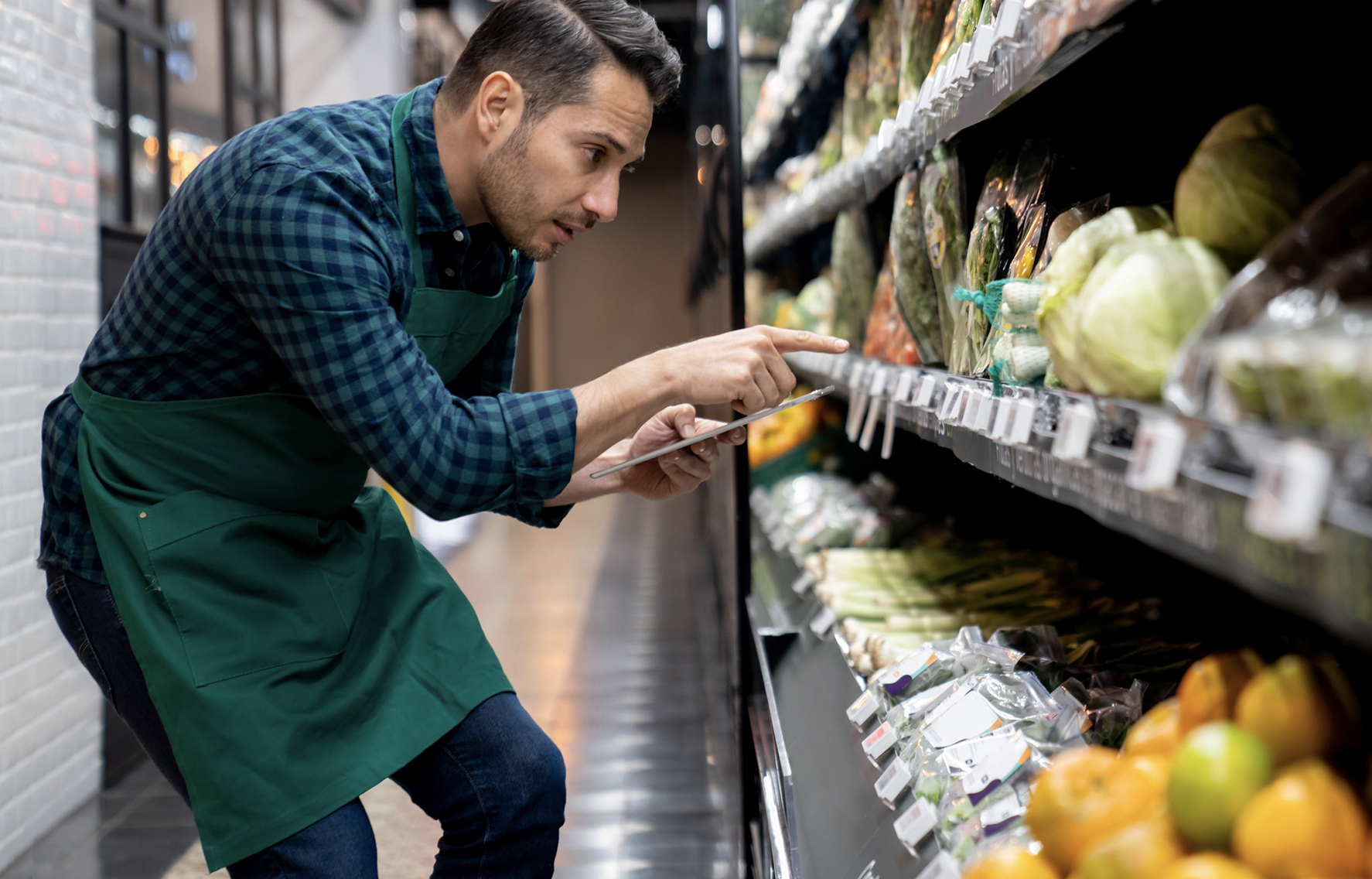 Grocery store employee kneeling and inspecting product labels on a grocery store shelf in the produce section. He is holding a clipboard and pointing at packaged vegetables.