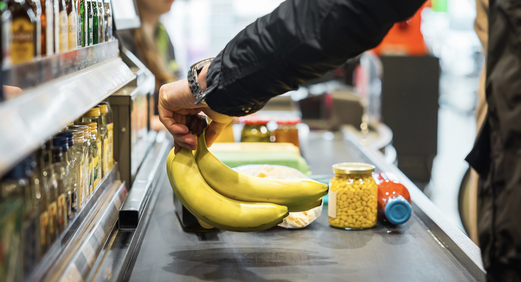 Close-up of a customer placing a bunch of bananas on a grocery store checkout conveyor belt with other food items.