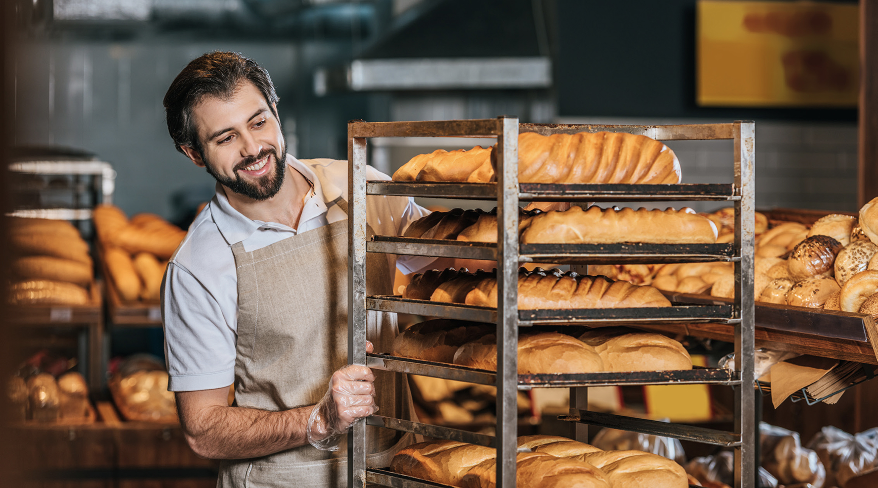 Man wearing a beige apron and clear plastic gloves smiling while pushing a metal rack filled with trays of freshly baked bread in a bakery setting.
