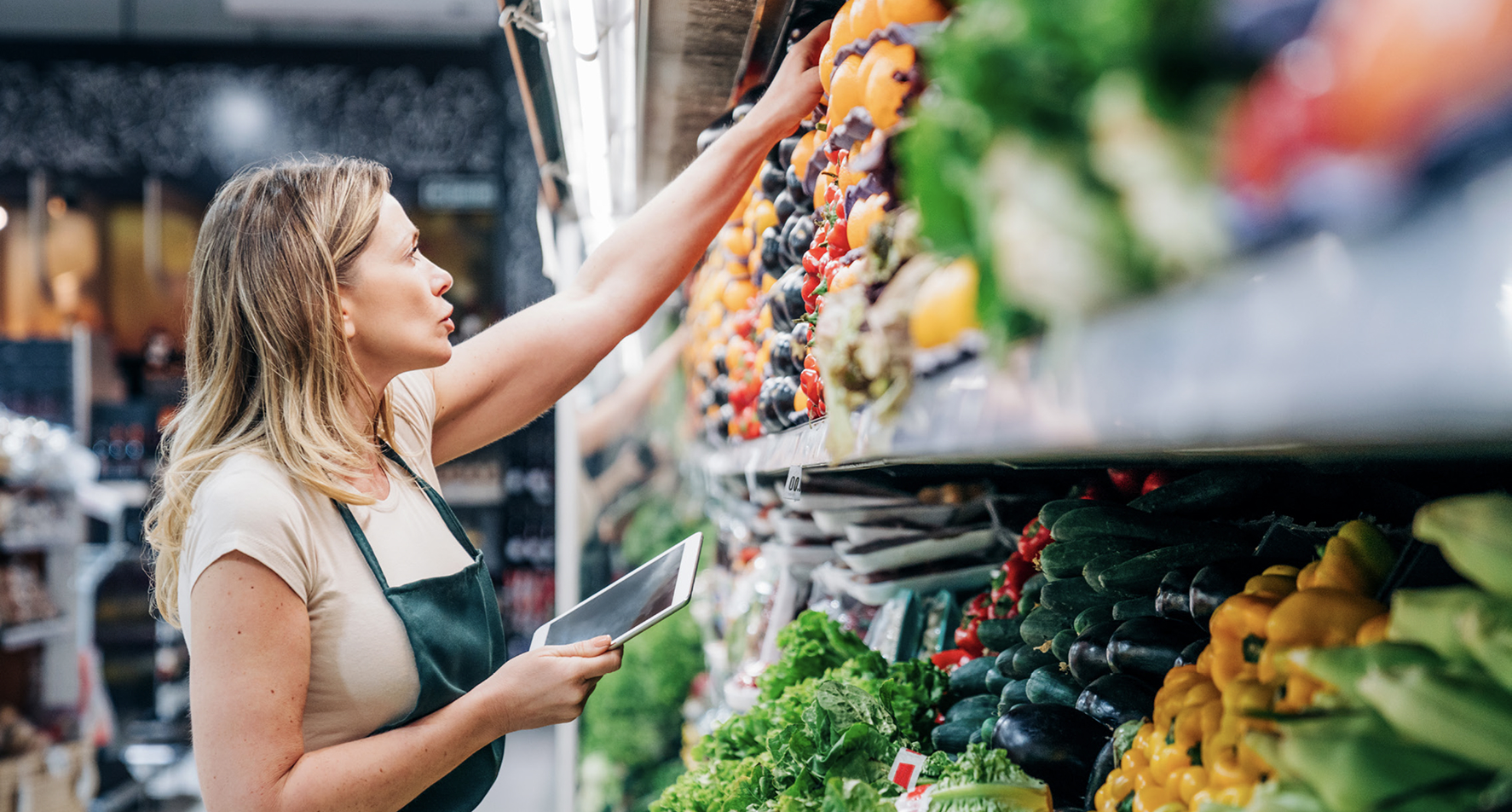 Female employee in a grocery store holding a tablet, reaching to stock or inspect produce on a shelf.
