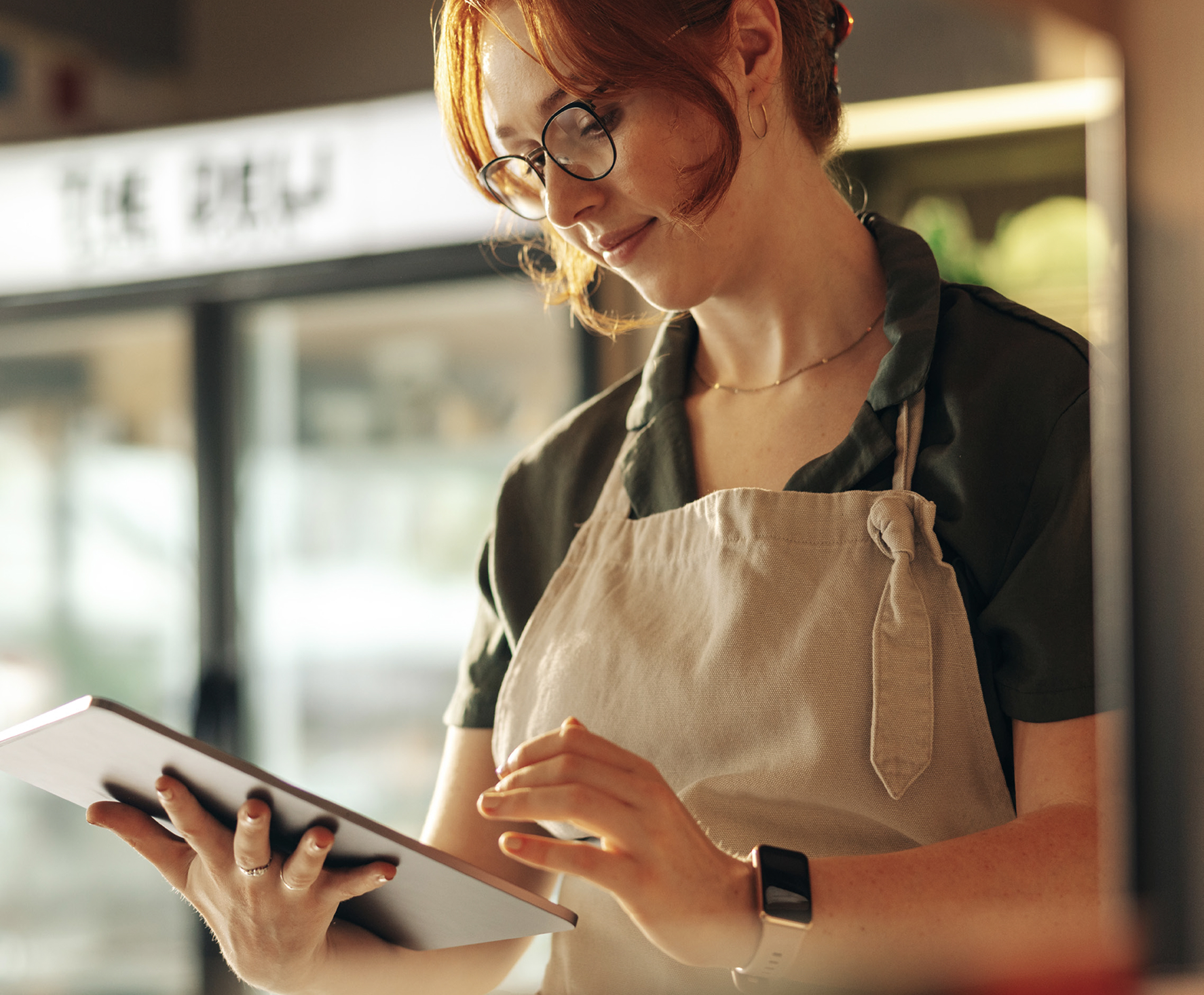 Woman with red hair and glasses wearing a beige apron and green shirt, using a tablet in a grocery or deli environment.