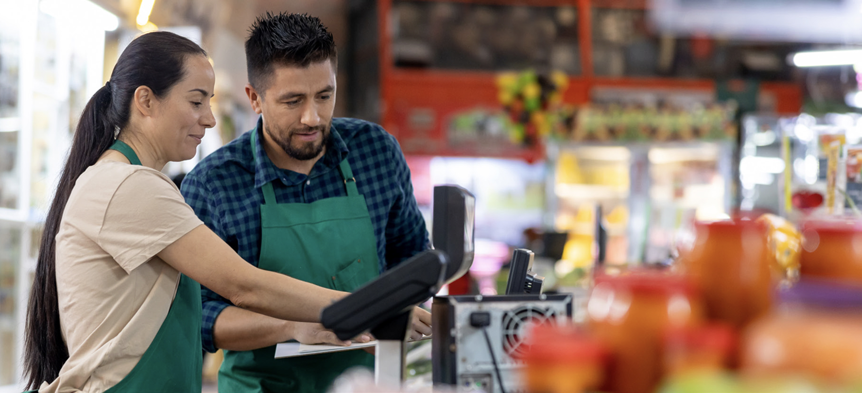 Two grocery store employees wearing green aprons are standing at a checkout station. The woman is pointing at the screen while the man observes, both appearing focused and engaged.