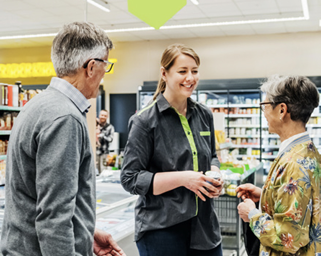Female grocery employee talking with two older customers in an aisle, smiling and holding a product can.