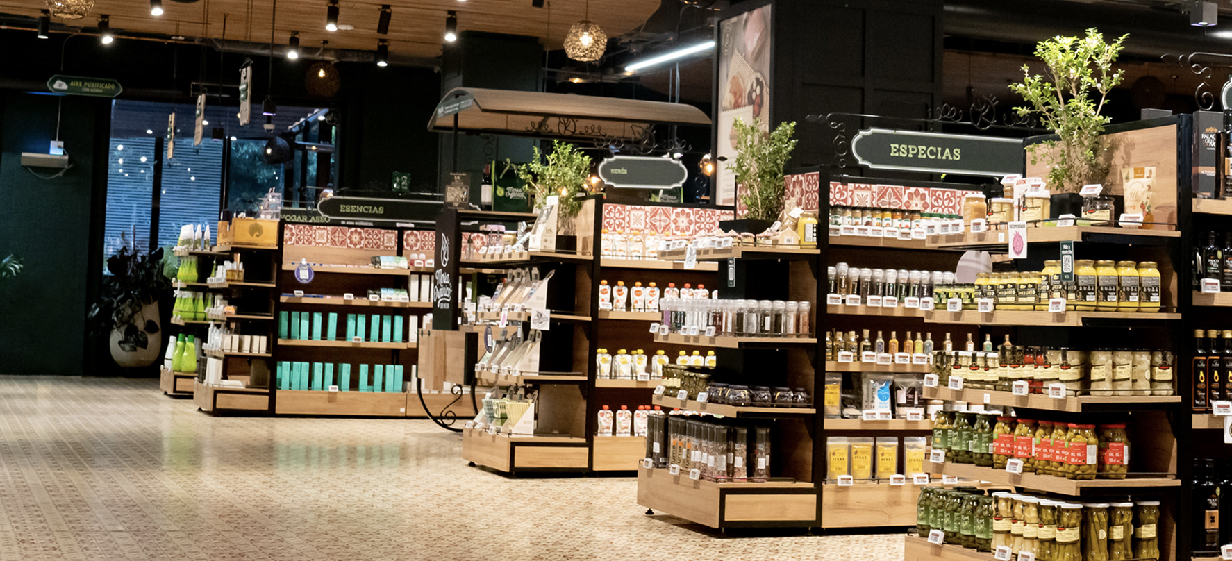 Wide-angle view of a modern grocery store interior with organized shelves labeled “Especias” and “Esencias” under warm lighting.