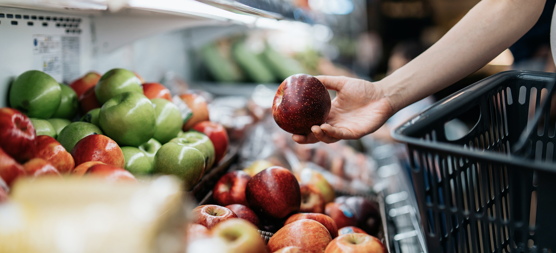 Close-up of a person holding a red apple while shopping in a grocery store produce section, with a black basket in hand.