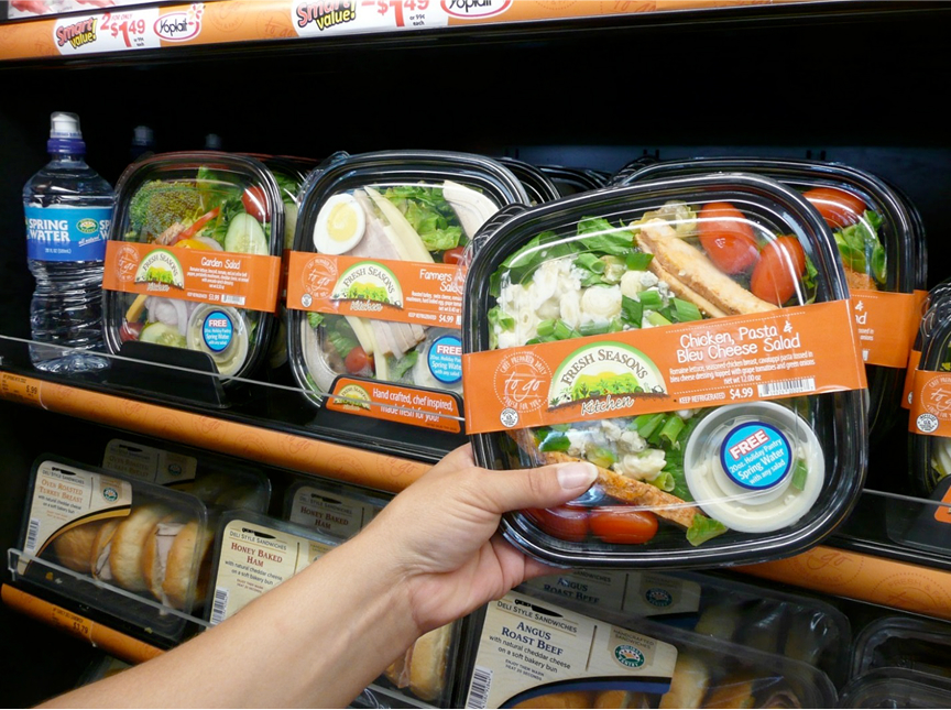  Image showing a refrigerated grocery shelf stocked with prepackaged salads and sandwiches. A hand is selecting a "Chicken, Pasta & Bleu Cheese Salad" by Fresh Seasons Kitchen, which includes a promotion for a free bottle of spring water with purchase. Other varieties visible include Garden Salad and Farmers Salad. Bottled water and deli sandwiches such as honey baked ham and roast beef are also on the shelf. The display emphasizes convenience and fresh, ready-to-eat meal options.