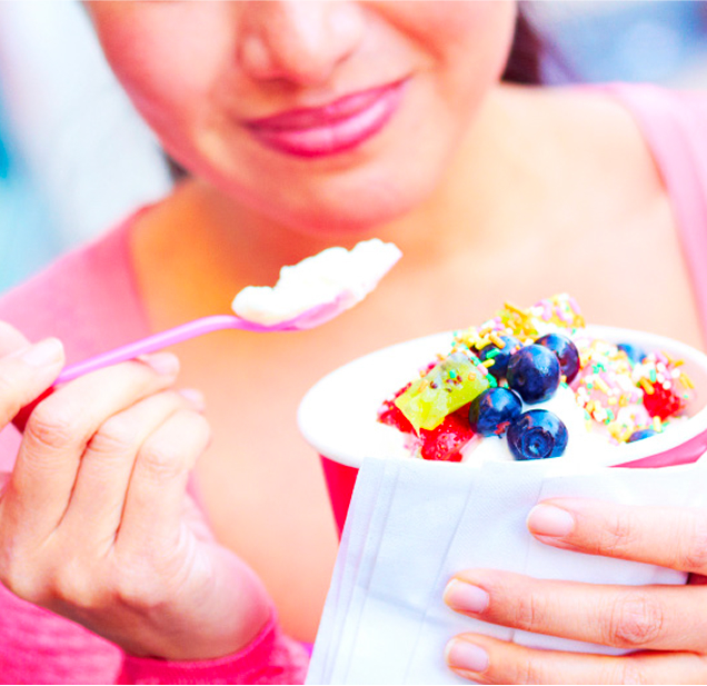 A woman holding a cup of frozen yogurt topped with fruits, gummy candies, and rainbow sprinkles. She is using a spoon to take a bite.