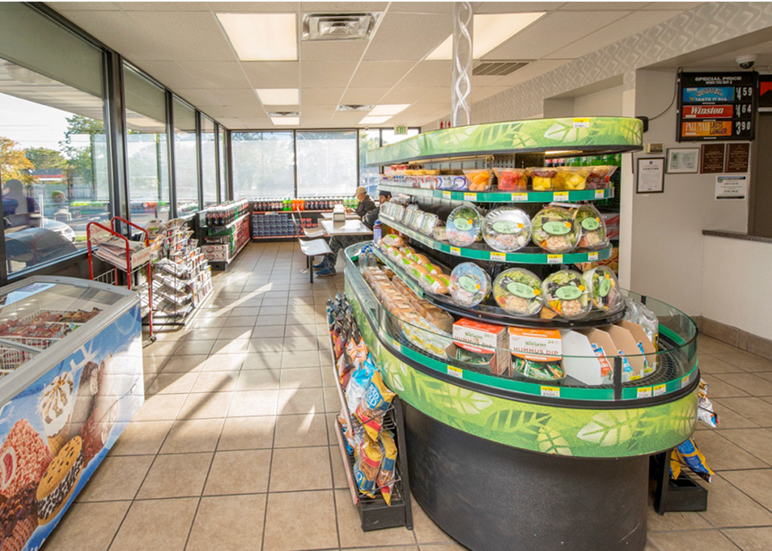 Interior of a convenience store with large front windows, tile flooring, and bright lighting. A refrigerated section displays fresh food items like salads and fruit cups. A seating area with tables is visible in the back. Freezers along the left wall contain frozen desserts. Shelves hold chips and beverages.