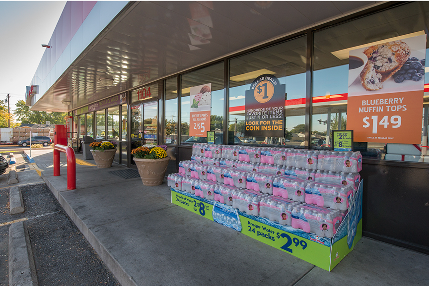  Exterior of a Loaf 'N Jug convenience store with promotional signs in the windows. Stacks of bottled water are displayed for sale in front of the store, but do not block any windows. Large potted flower arrangements are positioned on either side of the front door.