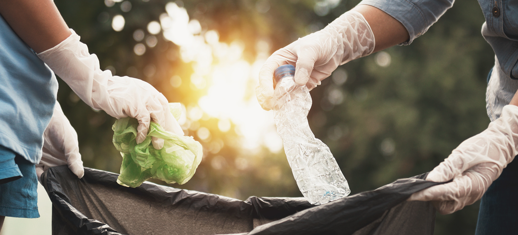 Two people wearing disposable gloves place a green plastic bag and a crushed plastic bottle into a black trash bag outdoors.