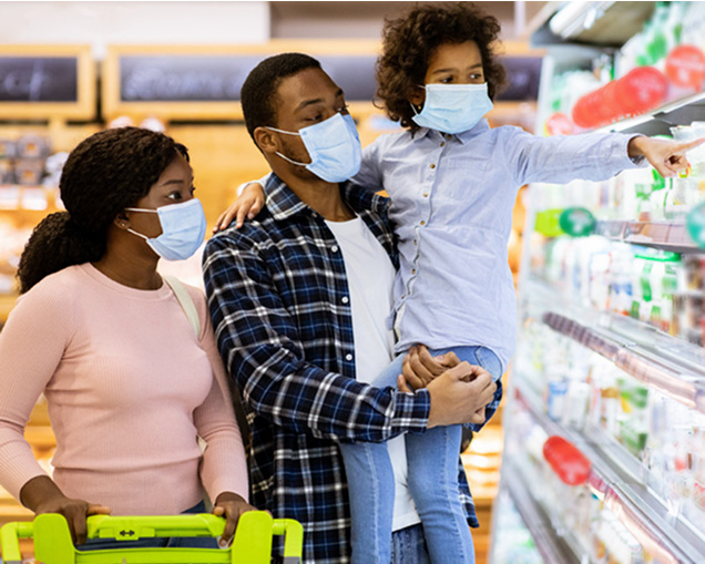 Family of three wearing face masks grocery shopping. The mother pushes a cart, the father holds a child who is pointing at items on a refrigerated store shelf.