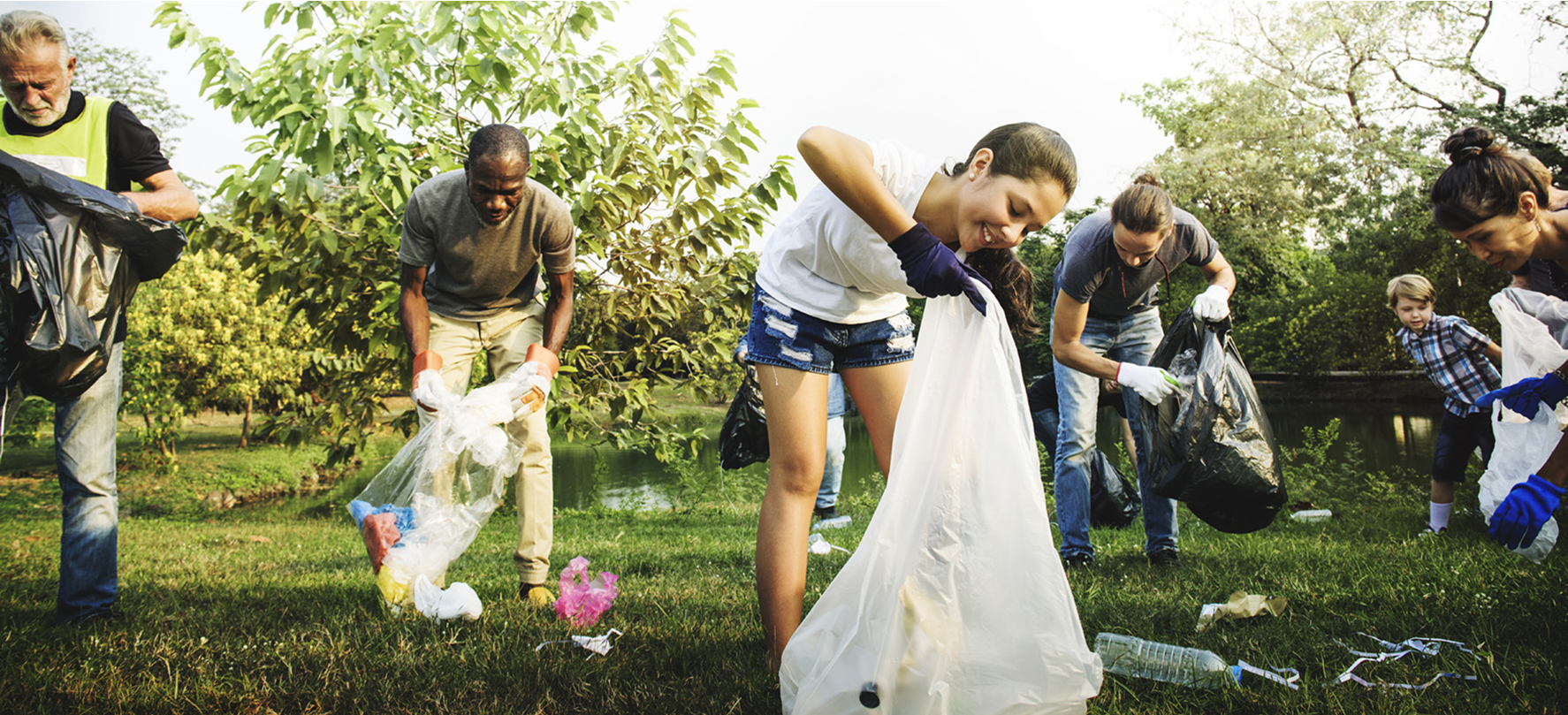 Group of people of various ages outdoors collecting litter in a grassy park area near a pond. They are wearing gloves and holding large trash bags while picking up plastic bottles and other debris.