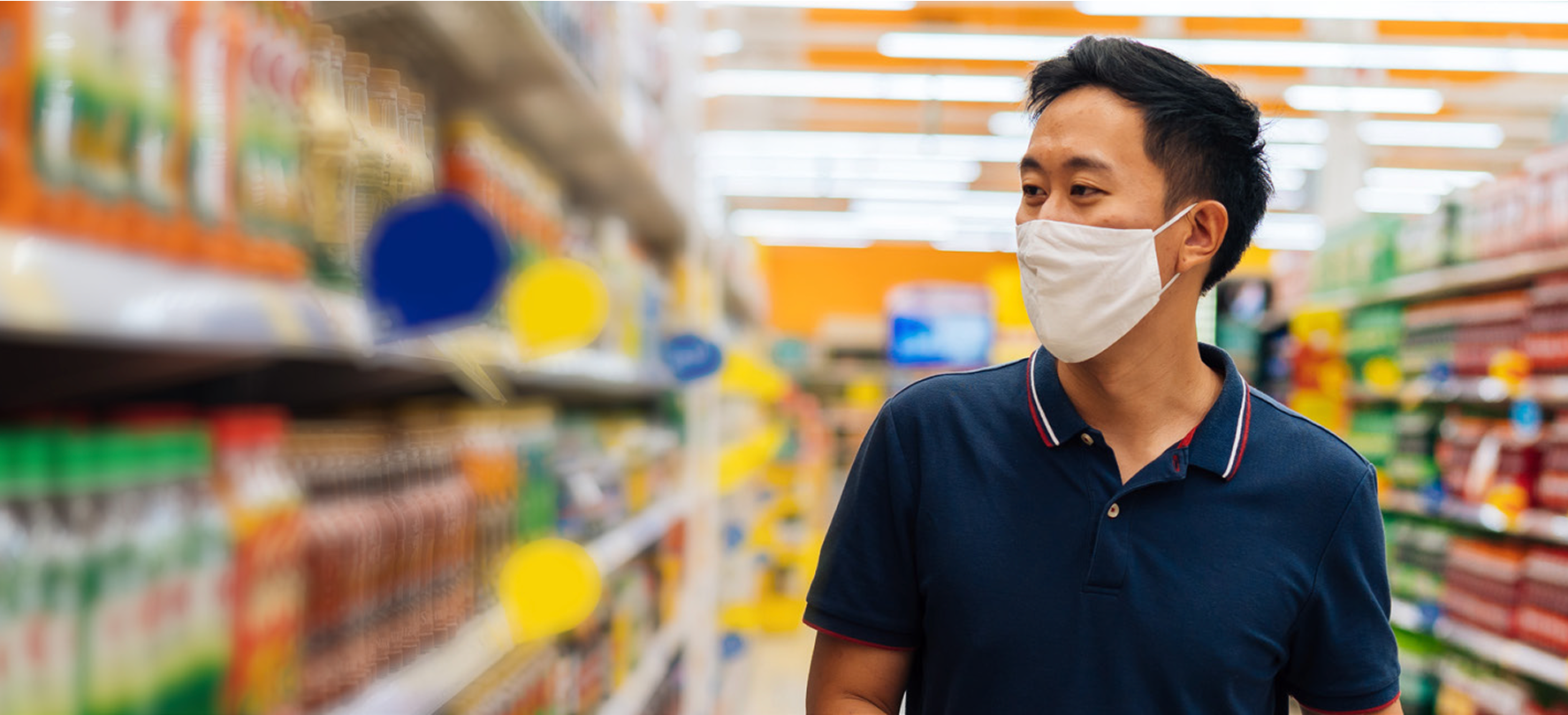 Man wearing a face mask walks through a brightly lit grocery store aisle lined with colorful bottled drinks.